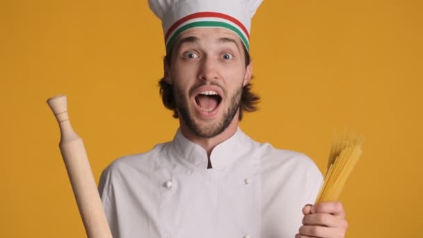Retrato del guapo chef italiano en uniforme sosteniendo el rodillo y los macarrones sonriendo alegremente en la cámara sobre un fondo colorido - Imágenes, Vídeo