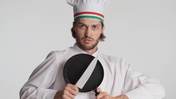 Attraktiver italienischer Koch in Uniform, der vor weißem Hintergrund mit Messer und Pfanne spielt. Herumalbern beim Ausdruck - Filmmaterial, Video