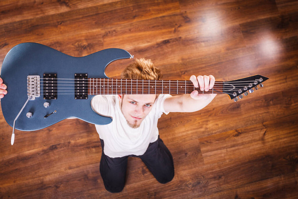 Young bearded man holding, raising his electric guitar. Hobby, music concept, on wooden floor. View from top - Foto, Bild