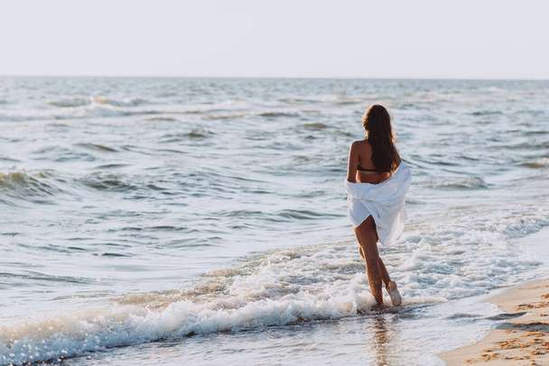 Young beautiful slender woman in a black bikini and white shirt on a tanned body on the beach in the waves, back view. Soft selective focus. - Foto, Bild