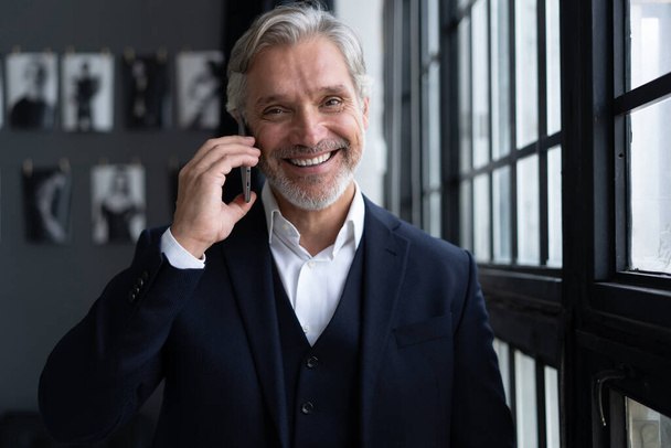 Success concept. Stylish mature bearded man in elegant suit talking on smartphone, standing near the window. - Photo, image