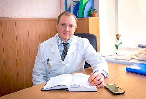 Jeune thérapeute caucasien médecin avec un sourire dans un manteau blanc assis dans son bureau attendant les patients. Médecine et santé concept. - Photo, image