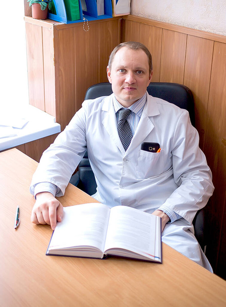 Ein junger kaukasischer Arzt mit einem Lächeln im weißen Mantel sitzt in seinem Büro und wartet auf Patienten. Medizin und Gesundheitskonzept. - Foto, Bild