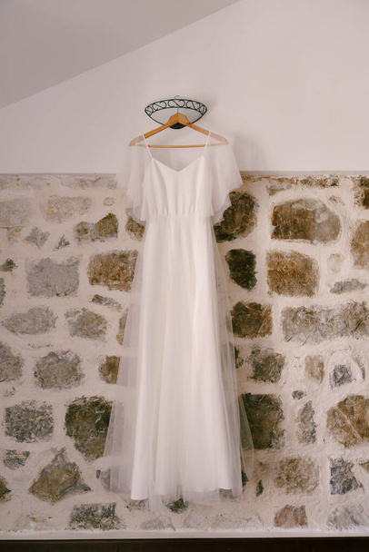 Vestido de novia blanco en una percha de madera contra una pared de piedra con un aplique en una habitación con un techo inclinado. - Foto, imagen