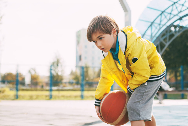 Ein süßer Junge in einer gelben Sportjacke wirft einen Basketball. Ein Kind spielt Basketball. Sport, Training, Lebensstil - Foto, Bild