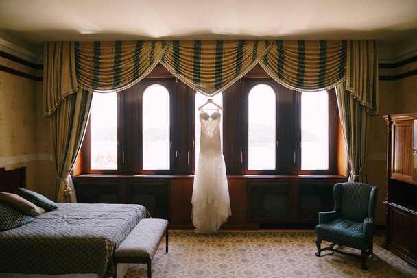 Lange witte jurk van de bruid op een hanger bij het raam in een kamer met een fauteuil en een bed. - Foto, afbeelding