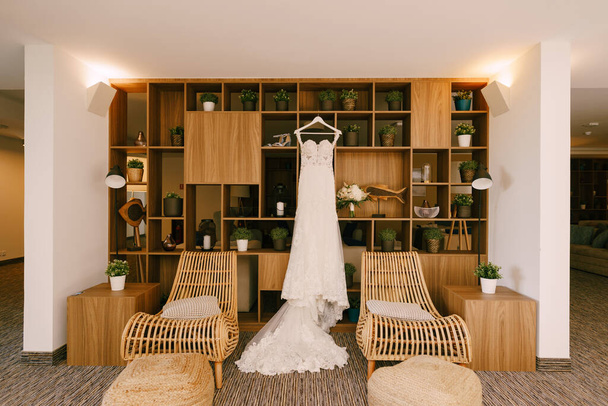 El vestido de novia con un tren largo en una habitación con sillones y un armario de madera con flores en macetas. - Foto, imagen