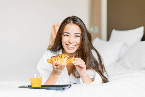 Hermosa sonriente joven mujer asiática con pelo largo en bata blanca desayunando croissant y jugo de naranja en la cama de la habitación de hotel - Foto, imagen