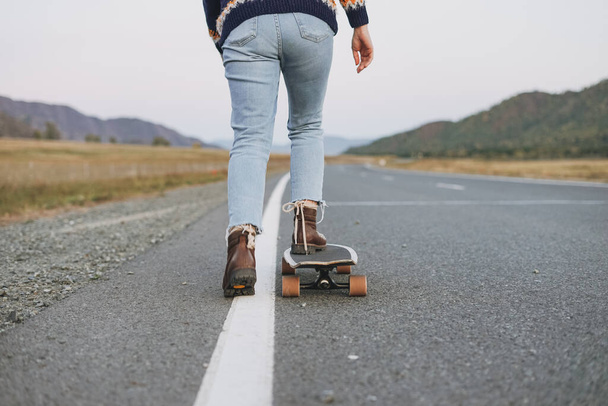 Schnappschuss von Frauenbeinen in Jeans auf Longboard auf der Straße mit Berghintergrund - Foto, Bild