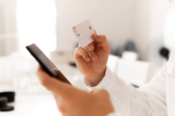 Mann reicht Kreditkarte für Online-Einkäufe. Die Hände eines Mannes, der eine Kreditkarte hält, Großaufnahme eines jungen Mannes, der im Internet einkauft. Online-Bezahlung, Zahlung Online-Shopping Smartphone am Arbeitsplatz. - Foto, Bild