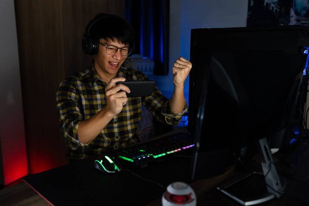 Izgalmas ázsiai férfi profi játékos ül az asztalnál, játszik és nyer online videojátékok egy számítógép és okostelefon, technológia játék cyber vagy e-sport bajnokság koncepció. - Fotó, kép