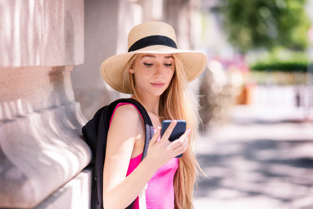 Shot of smiling young woman wearing straw hat and backpack while standing on the street and text messaging. - Photo, Image