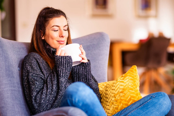 Portait of smiling woman relaxing in the armchair at home and drinking tea. - Foto, Imagem