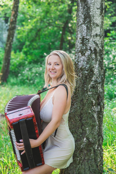 mooi blond in een nachtjapon speelt accordeon in het bos - Foto, afbeelding