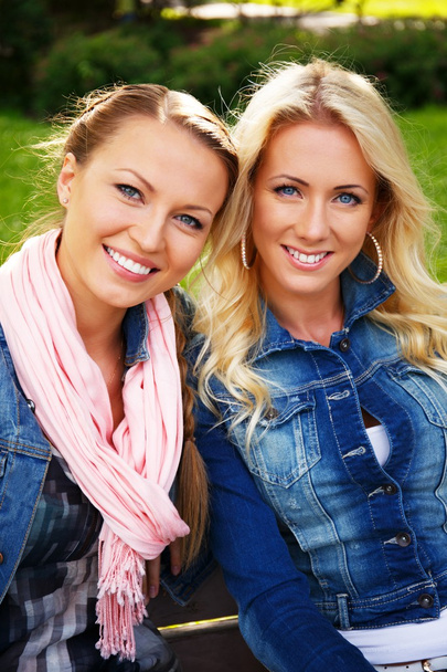 δύο όμορφες χαμογελώντας νεαρές γυναίκες με σακάκια τζιν που κάθεται σε ένα παγκάκι σε ένα πάρκο - Φωτογραφία, εικόνα