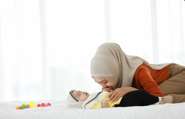  Η μητέρα φοράει μαντήλι και είναι ξαπλωμένη στο κρεβάτι κοντά στο μωρό. Του χαμογελάει ενώ κοιμάται. - Φωτογραφία, εικόνα