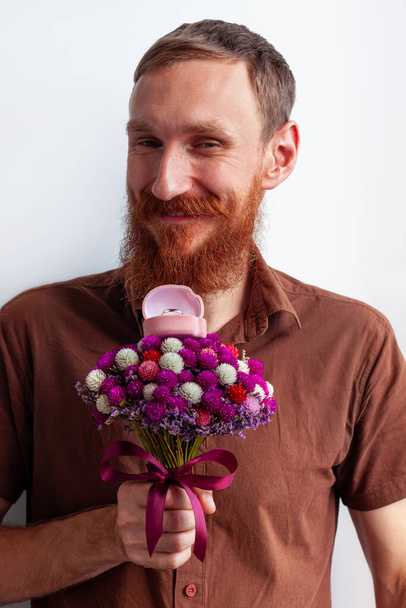 Marriage proposal. Handsome elegant man in brown shirt gives white gold ring in pink box beautiful flowers bouquet. Bearded guy proposing girlfriend to marry him. Romantic date, engagement celebration - Photo, Image