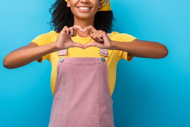 vue recadrée d'une femme afro-américaine joyeuse montrant un signe cardiaque avec les mains isolées sur bleu - Photo, image