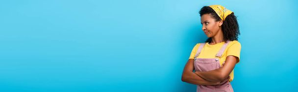 sceptyczna Afroamerykanka stojąca z skrzyżowanymi ramionami i odwracająca wzrok na niebieskim, sztandarze - Zdjęcie, obraz