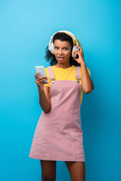 shocked african american woman in headphones listening music and using smartphone on blue - Fotó, kép