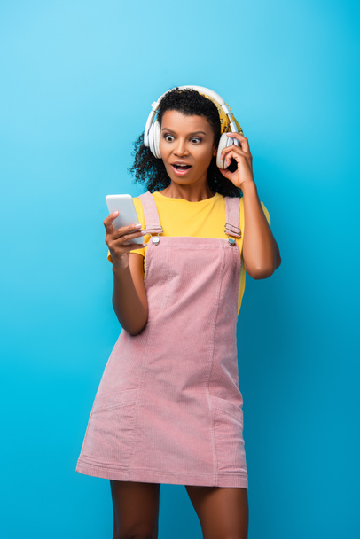 shocked african american woman in wireless headphones listening music and using smartphone on blue - 写真・画像