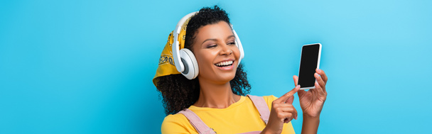 mujer afroamericana feliz en auriculares escuchando música y apuntando con el dedo al teléfono inteligente con pantalla en blanco en azul, bandera - Foto, imagen
