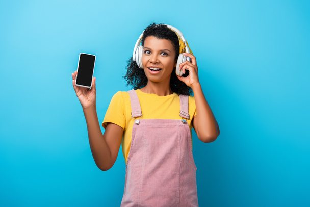 έκπληκτη Αφροαμερικανή γυναίκα με ασύρματα ακουστικά ακούγοντας μουσική και κρατώντας smartphone με λευκή οθόνη στο μπλε - Φωτογραφία, εικόνα