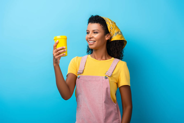 sonriente afroamericana mujer mirando reutilizable taza en azul - Foto, Imagen