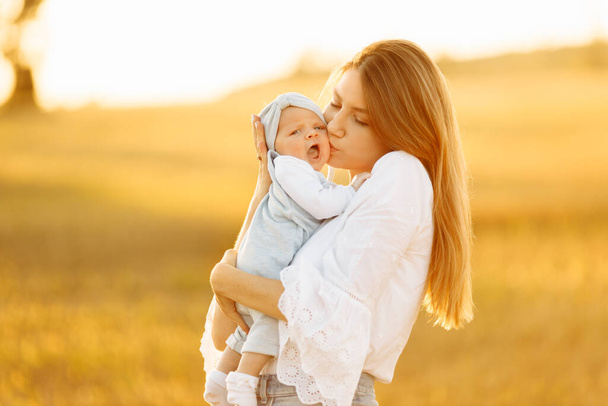 Adorable mother with beautiful little daughter at the field, pretty woman hold cute newborn baby in arms, caring mom gently kiss tiny baby girl, maternity concept   - Photo, image