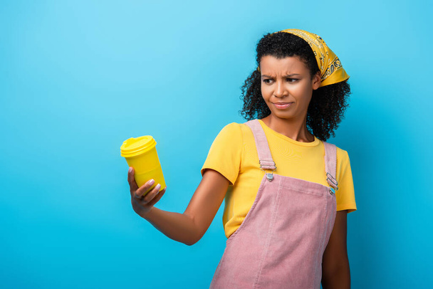 femme afro-américaine sceptique tenant tasse réutilisable sur bleu - Photo, image