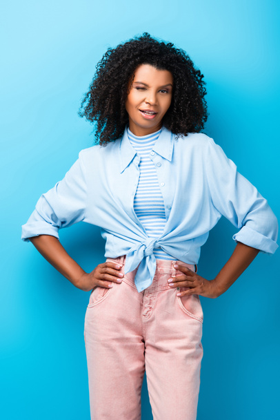 curly african american woman standing with hand on hips and winking eye on blue - Foto, immagini