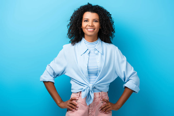 curly african american woman standing with hand on hips and smiling on blue - Foto, imagen