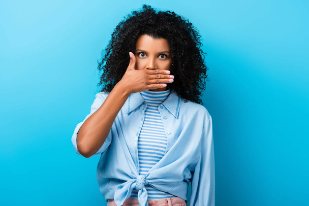 curly african american woman covering mouth on blue - Foto, Bild
