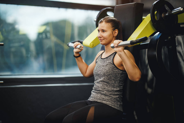 Shot of a muscular young woman in sportswear working out at the gym machine. She is pumping up her shoulder muscule with heavy weight. - Fotoğraf, Görsel