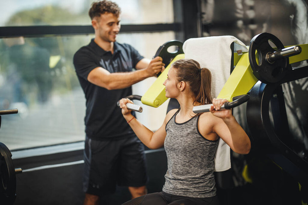 Shot of a muscular young woman in sportswear working out with personal trainer at the gym machine. She is pumping up her shoulder muscule with heavy weight. - Photo, Image