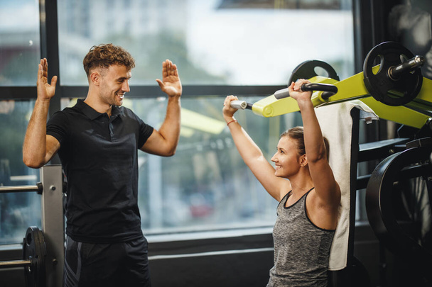 Foto de una joven musculosa en ropa deportiva haciendo ejercicio con un entrenador personal en la máquina de gimnasio. Ella está bombeando su músculo del hombro con peso pesado. - Foto, imagen