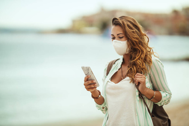 Aufnahme einer attraktiven, glücklichen jungen Frau, die eine schützende N95-Maske trägt und mit ihrem Smartphone in den sozialen Medien surft, während sie ihren Urlaub am Strand während der COVID-19 genießt. - Foto, Bild