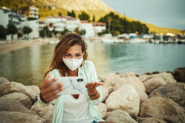 Aufnahme einer attraktiven, glücklichen jungen Frau, die eine schützende N95-Maske trägt und mit ihrem Smartphone Videotelefonate führt, während sie während der COVID-19 Zeit am Meer verbringt. - Foto, Bild