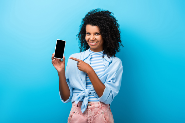 mulher americana africana feliz apontando com o dedo para o smartphone com tela em branco no azul - Foto, Imagem