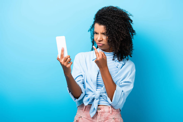 dissatisfied african american woman showing middle finger to smartphone on blue - Фото, зображення