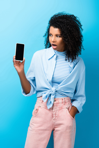 schockierte Afroamerikanerin steht mit der Hand in der Tasche und hält Smartphone auf blauem Grund - Foto, Bild