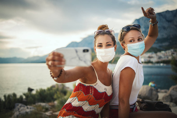 Shot of attractive female friends wearing with protective mask and making selfie with smartphone while exploring the wonderful city of Mediterranean. - Photo, Image