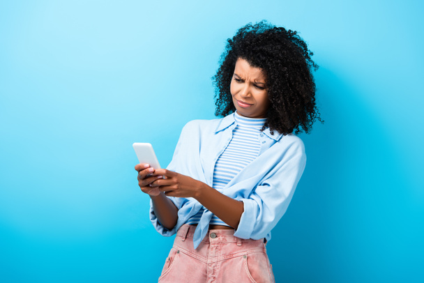displeased african american woman texting on smartphone on blue - Фото, изображение