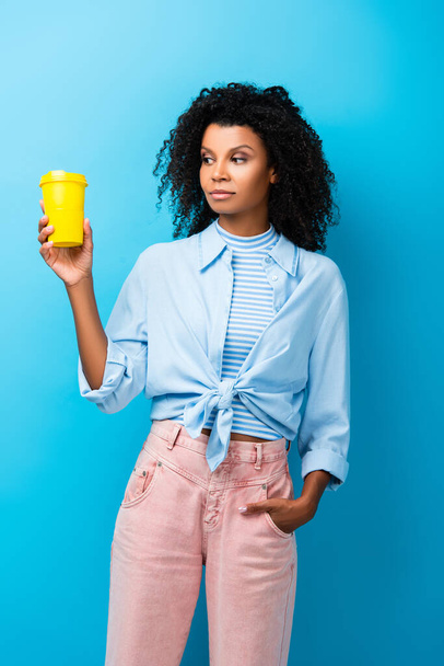 mujer afroamericana sosteniendo taza reutilizable vacía mientras está de pie con la mano en el bolsillo en azul - Foto, imagen