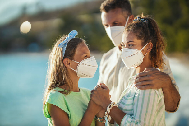 Foto de una familia feliz abrazado con máscara protectora N95 pasar tiempo en la playa en pandemia corona. Están teniendo auto-aislamiento en la naturaleza. - Foto, Imagen