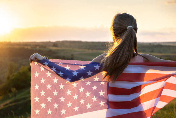 Onnellinen nuori nainen poseeraa USA:n lipun kanssa ulkona auringonlaskun aikaan. Positiivinen tyttö juhlimassa Yhdysvaltain itsenäisyyspäivää. Kansainvälinen demokratian päivä käsite. - Valokuva, kuva