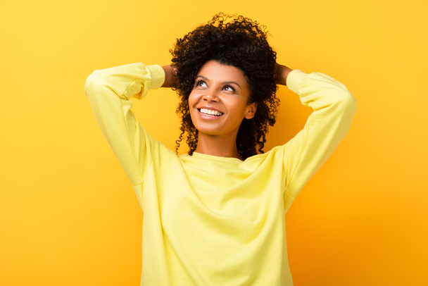 šťastný africký americký žena dívá pryč při upevňování kudrnaté vlasy na žluté - Fotografie, Obrázek