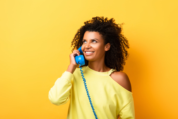 alegre afroamericana mujer hablando en vintage teléfono en amarillo - Foto, Imagen