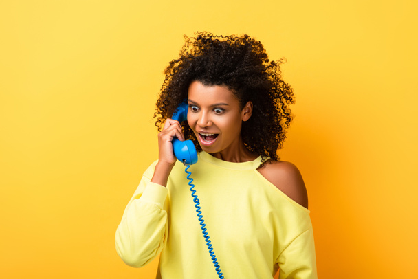 šokovaný africký Američan žena mluví na vintage telefon na žluté - Fotografie, Obrázek