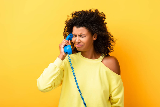 upset african american woman talking on vintage telephone on yellow - Photo, Image